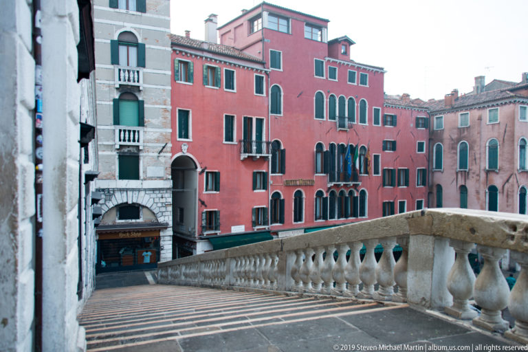 Stone landing and balustrade on the Rialto Bridge facing red and pale Venetian buildings with green shutters.