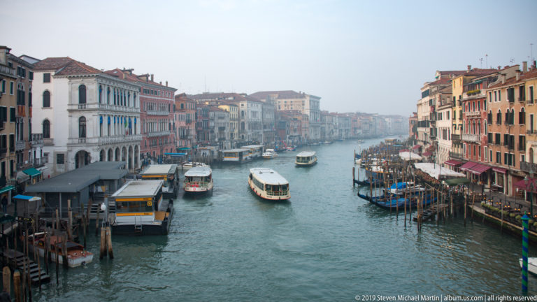 Elevated view from the Rialto Bridge along the Grand Canal, with vaporetti, docks, palazzi, and busy waterfronts.