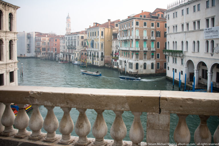 Grand Canal and rows of palazzi seen over the pale stone balustrade of the Rialto Bridge.