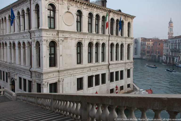 Pale Renaissance palazzo beside the Rialto Bridge with arched windows, pilasters, flags, and a canal view beyond.