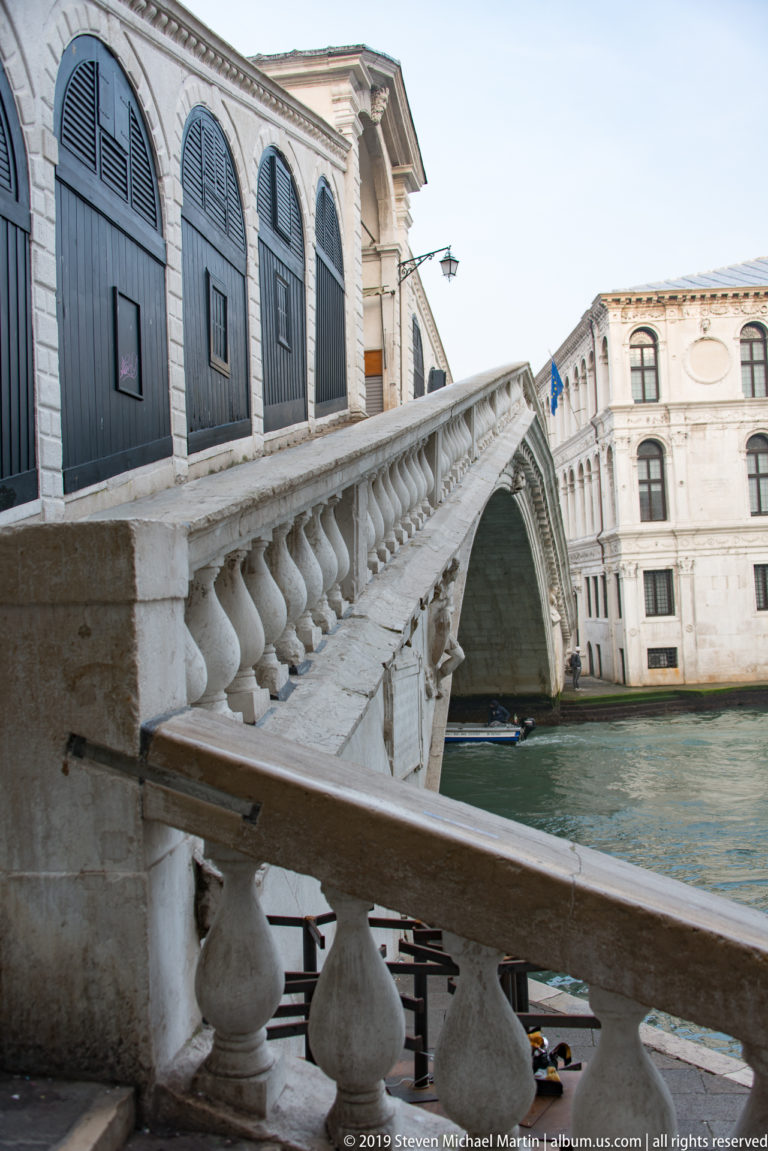 Weathered stone balustrade on the Rialto Bridge leading toward its arch, with shuttered shops along the left.