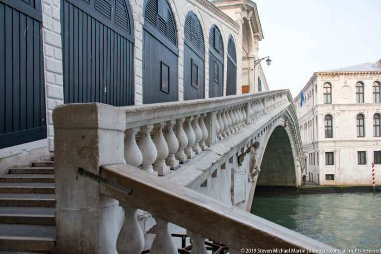 Stone stair and repeating balusters along the Rialto Bridge, with dark shop shutters and the canal beyond.