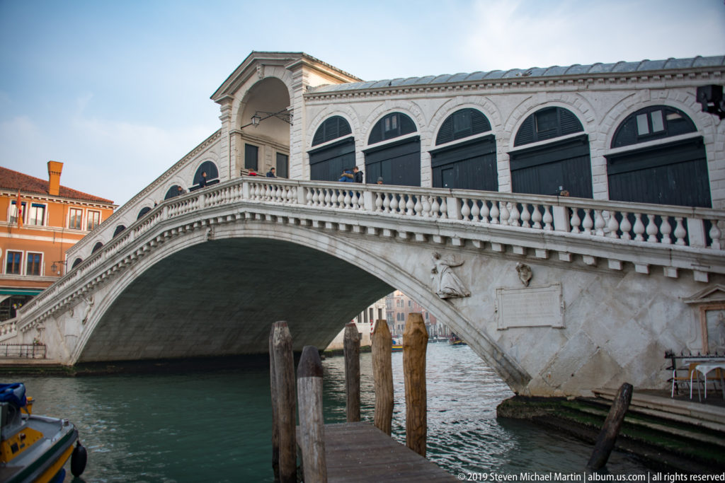 Side view of the Rialto Bridge's broad stone arch, balustraded ramp, and shop openings above the Grand Canal.