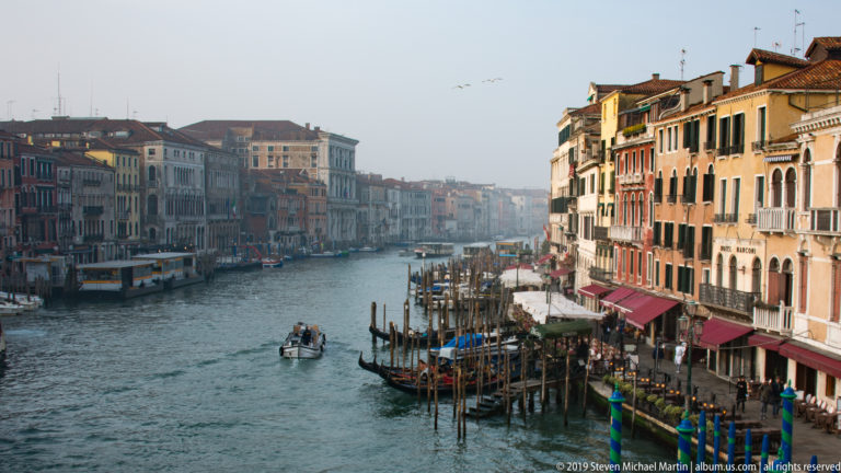 Grand Canal seen from the Rialto Bridge with a motorboat, gondola moorings, striped poles, and palazzi along both banks.