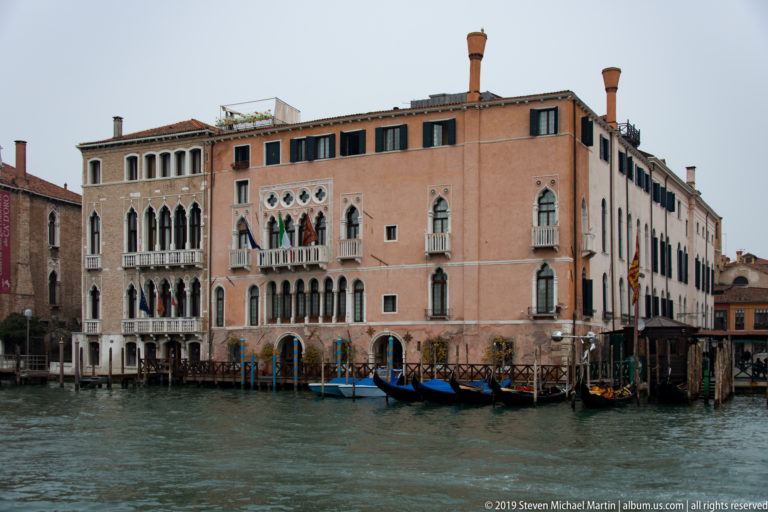 Salmon-colored Venetian palace with Gothic windows and balconies rises directly from the Grand Canal beside gondolas and timber mooring posts.