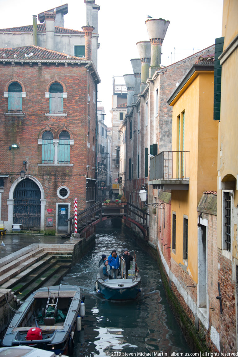 Small motorboat traveling along a narrow canal between brick and stucco buildings, with a low bridge ahead.
