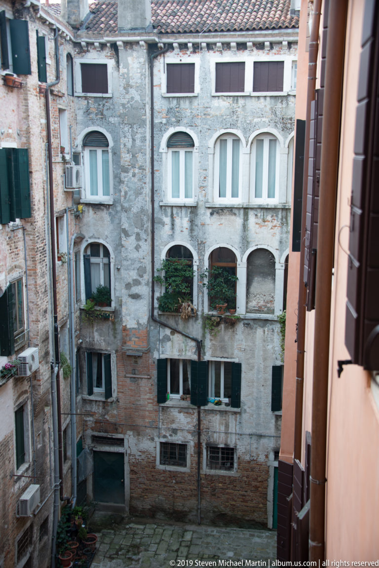 Weathered courtyard façade with exposed brick, gray plaster, green shutters, flower boxes, and irregular windows.