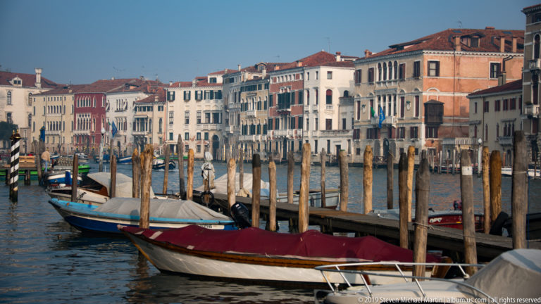 Small boats moored among many timber posts on the Grand Canal, backed by rows of pastel Venetian palazzi.