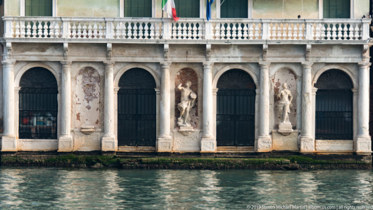 Marble waterfront façade with tall arched openings, recessed statues, a balustraded upper level, and flags above the canal.