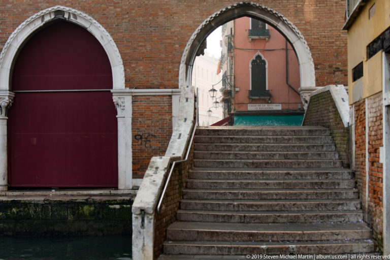 Stone steps rise between balustrades to a pointed brick arch framing a narrow Venetian street beyond.