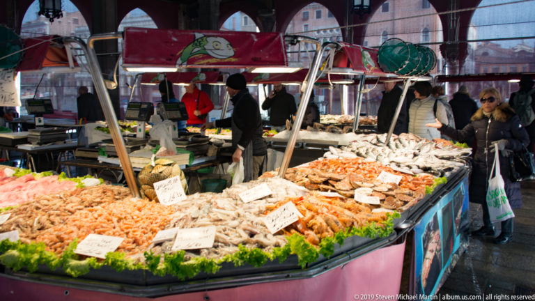 Curved seafood counter piled with shrimp, fish, and shellfish beneath hanging lamps, with vendors and shoppers behind.