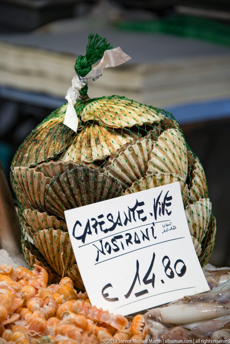 Woven basket of scallops at a market counter with a handwritten 'Capesante Nostrane' sign priced at €14.80.