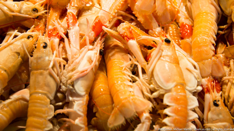 Close-up of orange-pink langoustines piled together, showing claws, segmented tails, black eyes, and long antennae.
