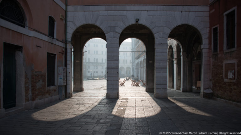 Three round stone arches at the end of a shaded passage opening onto a bright square with distant pedestrians.
