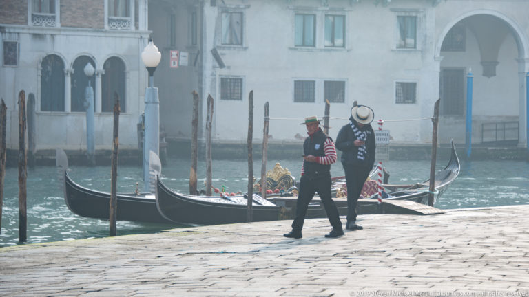 Two gondoliers in striped shirts and straw hats walking past moored gondolas on a bright stone waterfront.
