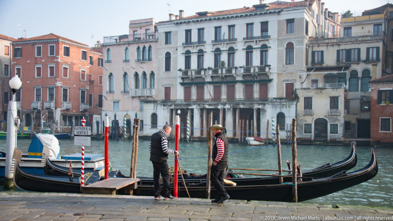 Two gondoliers beside moored black gondolas and red-and-white poles, with Grand Canal residences across the water.