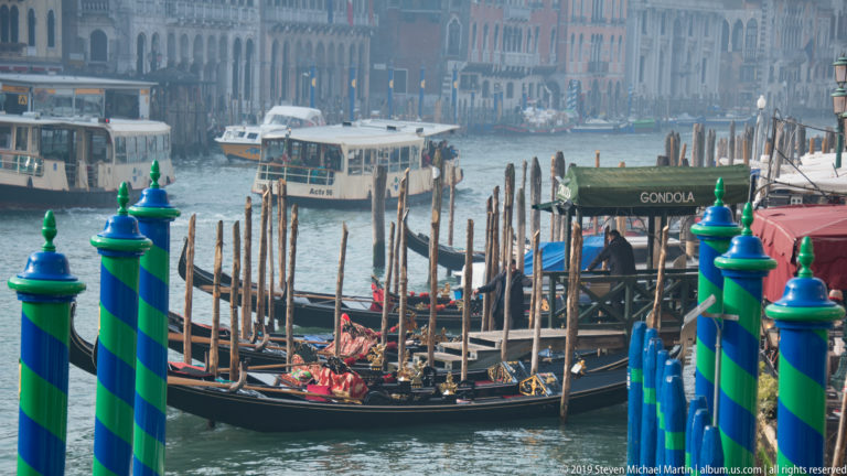 Green-and-blue striped poles and moored gondolas in the foreground, with public water buses crossing the Grand Canal behind.