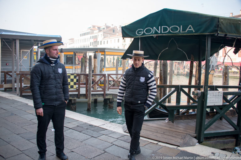 Two gondoliers in striped shirts and dark vests standing beside a covered gondola station on the waterfront.