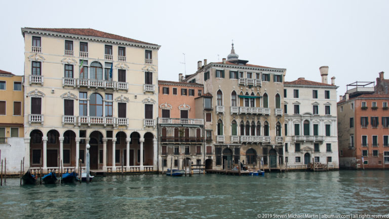 Row of ornate Grand Canal palazzi with arcades, balconies, Gothic windows, and pale façades reflected in the water.