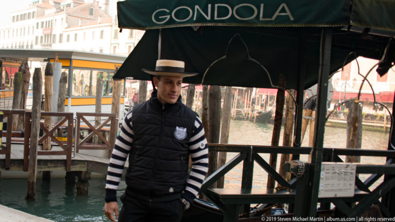 Gondolier in striped shirt and dark vest standing beneath a canopy labeled 'GONDOLA' beside a canal landing.