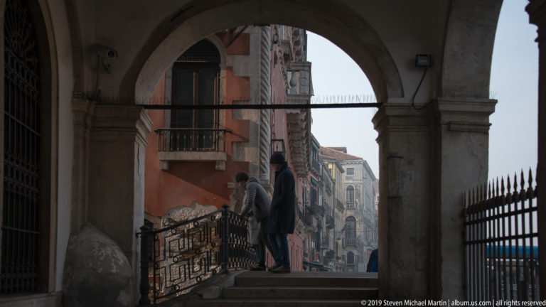 Large stone arch framing steps and pedestrians on a Venetian street, with iron railings and historic façades beyond.