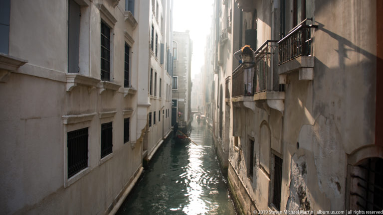 Backlit narrow Venetian canal between tall buildings, with a projecting balcony on the right and sunlight reflecting on the water.