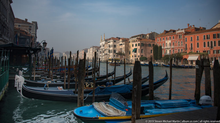 Several black gondolas and a blue motorboat moored among timber posts beside the Grand Canal, with palazzi opposite.