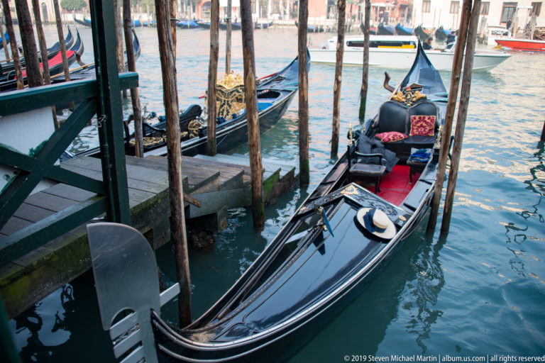 Black gondola moored between wooden posts, with upholstered seats, decorative textiles, and a straw gondolier hat aboard.