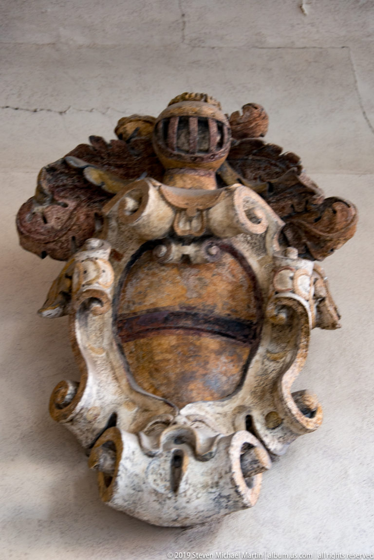 Carved heraldic shield with crest and scrolling Baroque surround, retaining traces of ochre, pink, and dark paint.