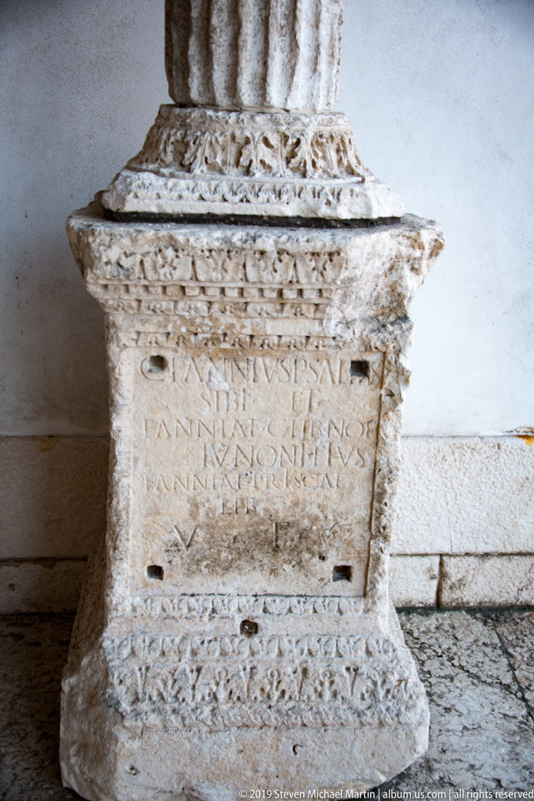 Weathered carved stone base with a Latin inscription and bands of foliate ornament, set against a pale wall.