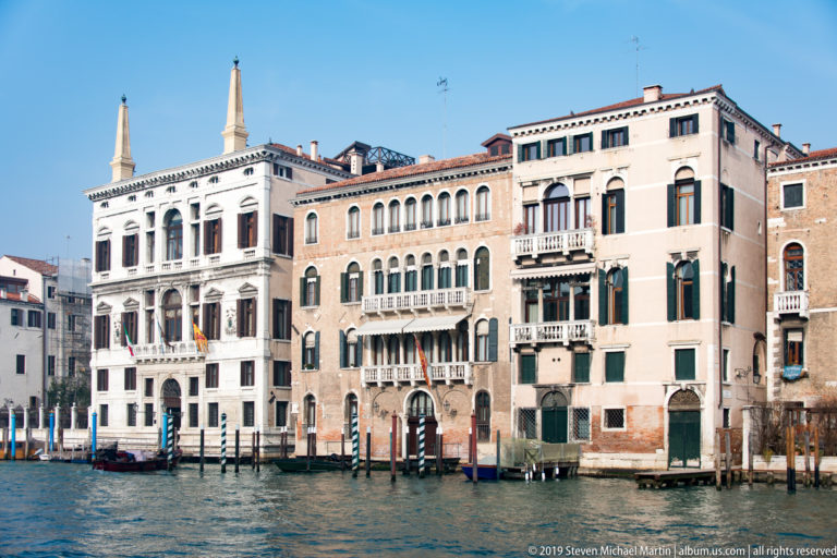 Three adjoining Grand Canal palazzi with arched windows, balconies, shutters, water doors, and mooring posts.