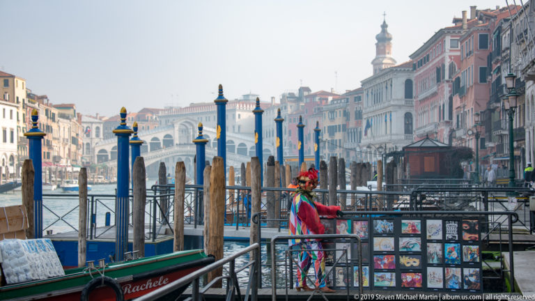 Rialto Bridge beyond gondolas, blue-and-gold mooring poles, timber posts, and a street vendor's display.