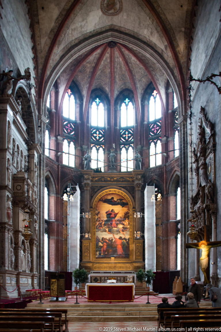 High altar and large altarpiece centered beneath a semicircle of tall Gothic apse windows, viewed from farther back in the church.