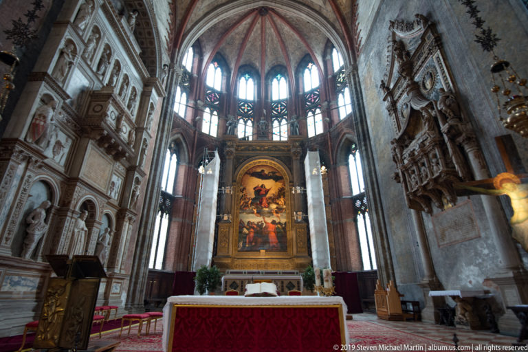 Wide sanctuary view with a red-draped altar, large high-altarpiece painting, tall Gothic windows, and monumental tombs on both side walls.
