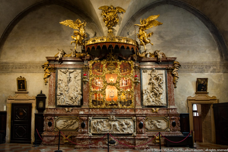 Large polychrome marble devotional structure with gilded angels, a richly decorated central panel, and white sculpted reliefs on both sides.