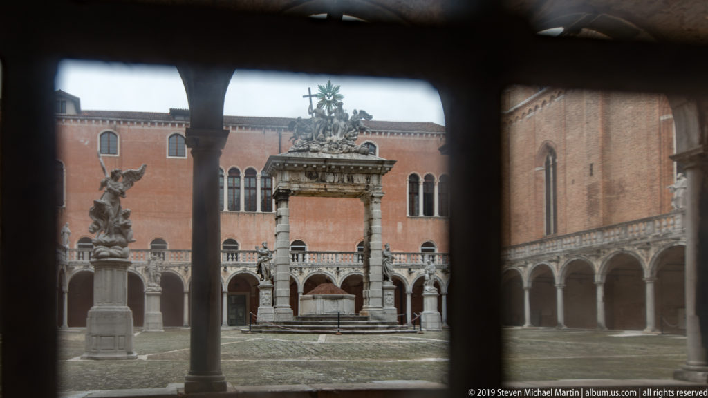 Sunlit cloister courtyard seen through dark columns, with stone monuments, palms, and arcaded brick buildings surrounding the open space.