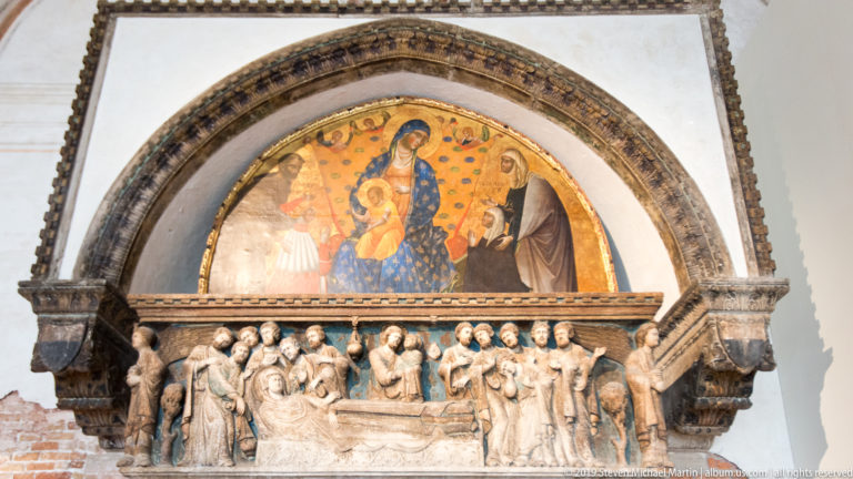Arched wall monument with a painted Madonna and Child above a carved relief of mourners gathered around a recumbent figure.