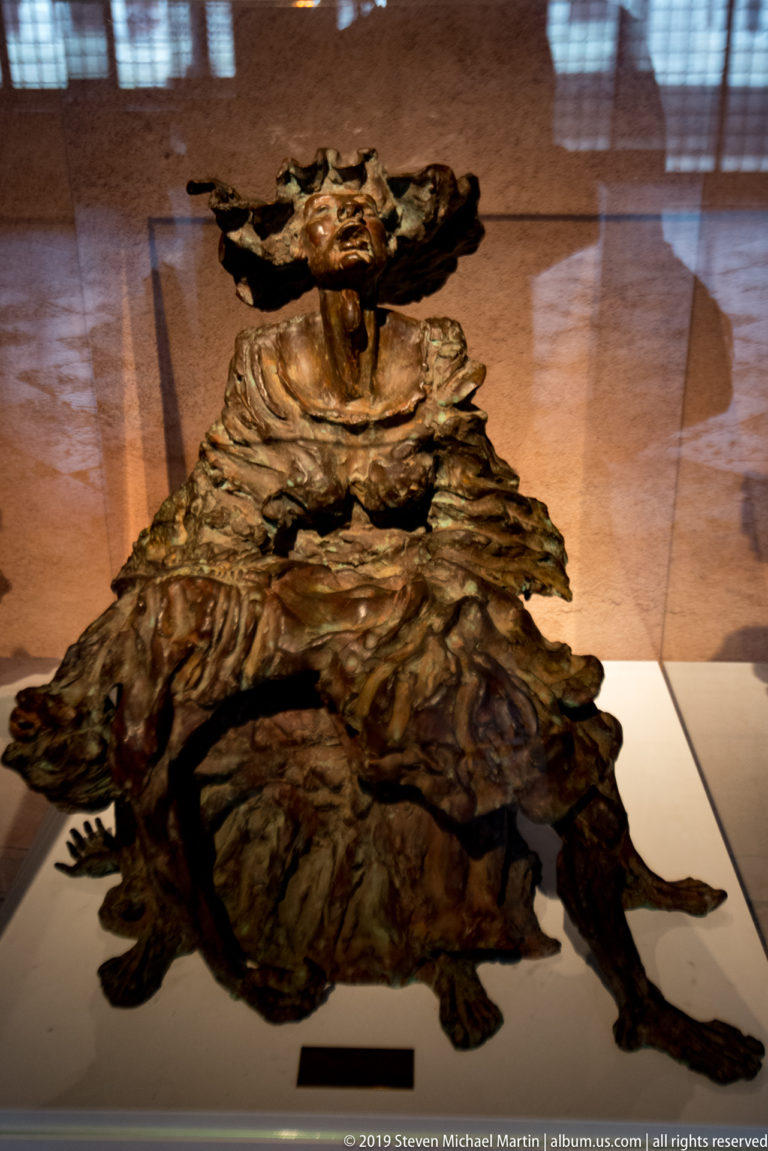 Expressive seated robed sculpture behind glass, with head turned upward and deeply modeled folds spreading across an irregular base.