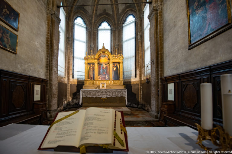 Open liturgical book on a foreground altar facing a gilded altarpiece between tall lancet windows in a narrow chapel.