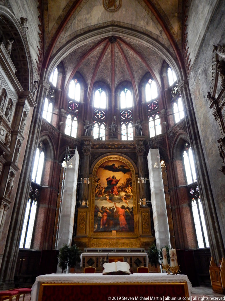 High altar with a large Assumption painting centered beneath tall Gothic apse windows, with altar table and floral arrangements in front.