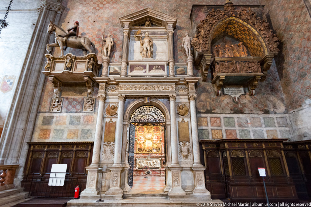 Elaborate marble portal with columns and statues beneath adjacent funerary monuments, including a projecting pointed-arch tomb at upper right.