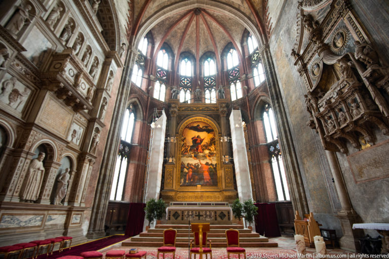 High altar and large painted altarpiece centered beneath tall Gothic apse windows, with monumental sculptural tombs on both side walls.