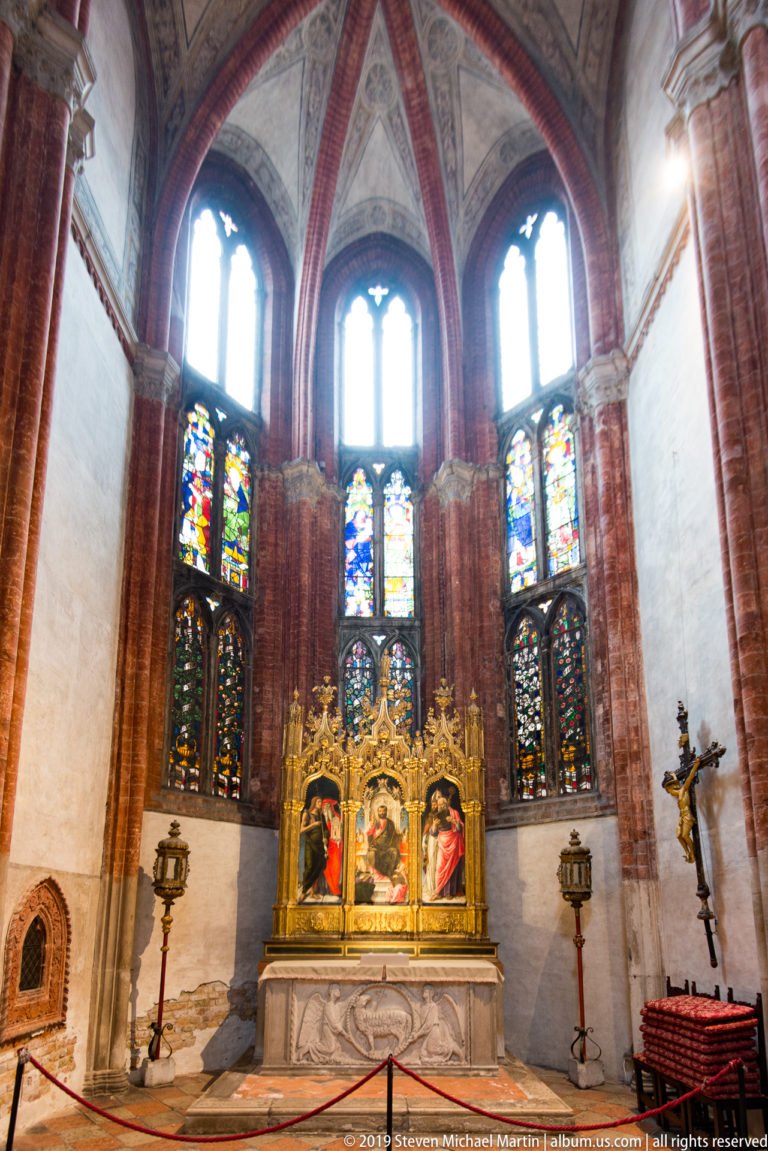 Wide view of a gilded triptych and altar beneath tall stained-glass windows in a ribbed red-brick Gothic chapel.