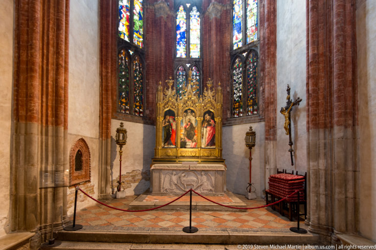 Gilded triptych on a stone altar beneath tall stained-glass windows, with a suspended crucifix and brick Gothic architecture nearby.