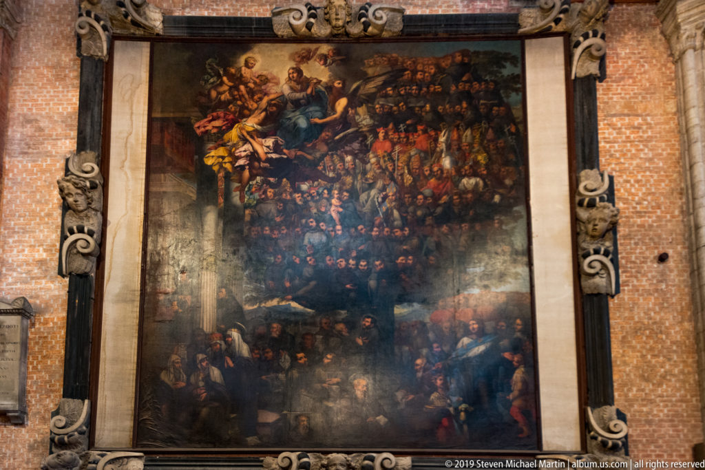 Large dark religious painting with many figures in an ornate vertical frame mounted against the basilica’s red-brick wall.
