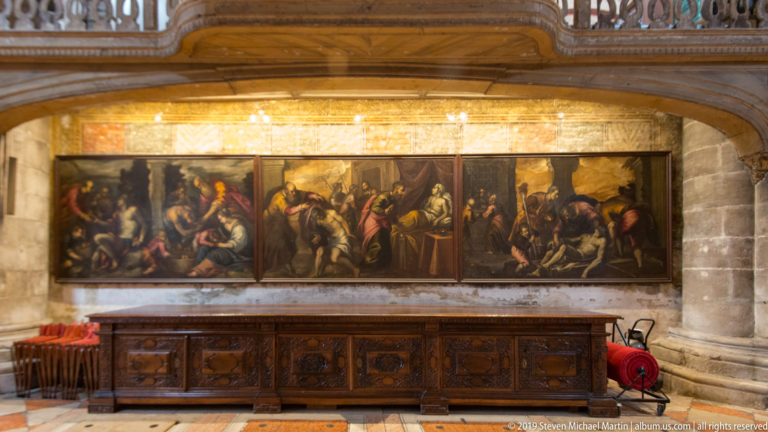 Three large dark religious paintings hang in a row above carved wooden furnishings beneath a broad stone arch inside the basilica.
