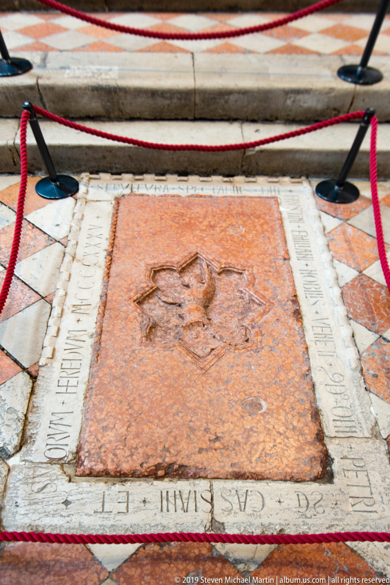 Reddish rectangular grave slab with a worn carved emblem and pale inscribed border set into a patterned stone floor.