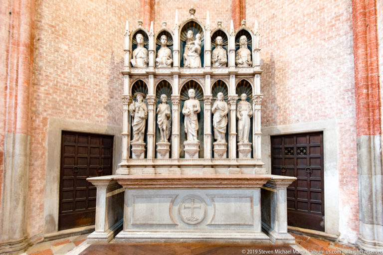 Two-tier Gothic stone altarpiece with standing saints in pointed niches, slender colonnettes, and pinnacles against a red-brick wall.