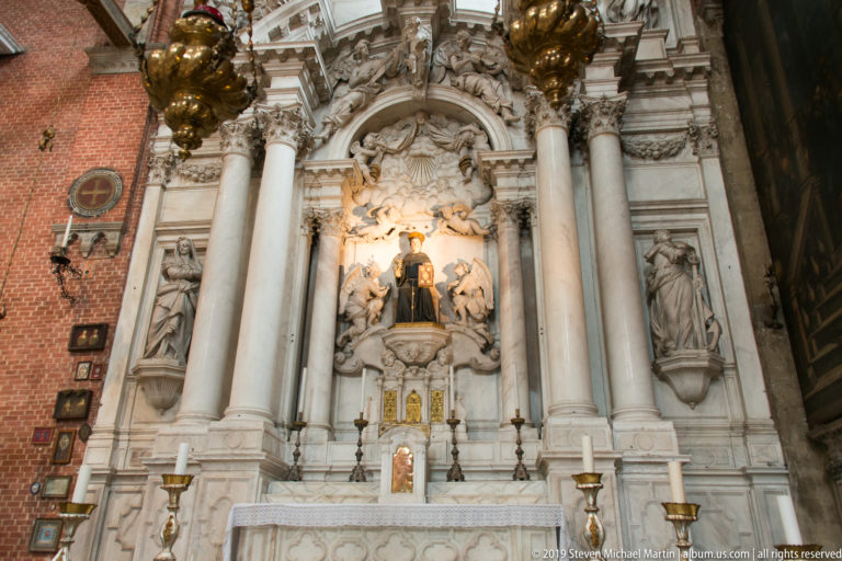 Full-height ornate side altar against red brick, with tall columns, a central dark-robed saint, angels, and additional sculpted figures.