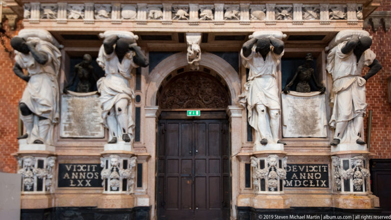 Dark central doorway framed by large draped supporting figures, niche statues, and black funerary inscription panels with Roman-numeral dates.